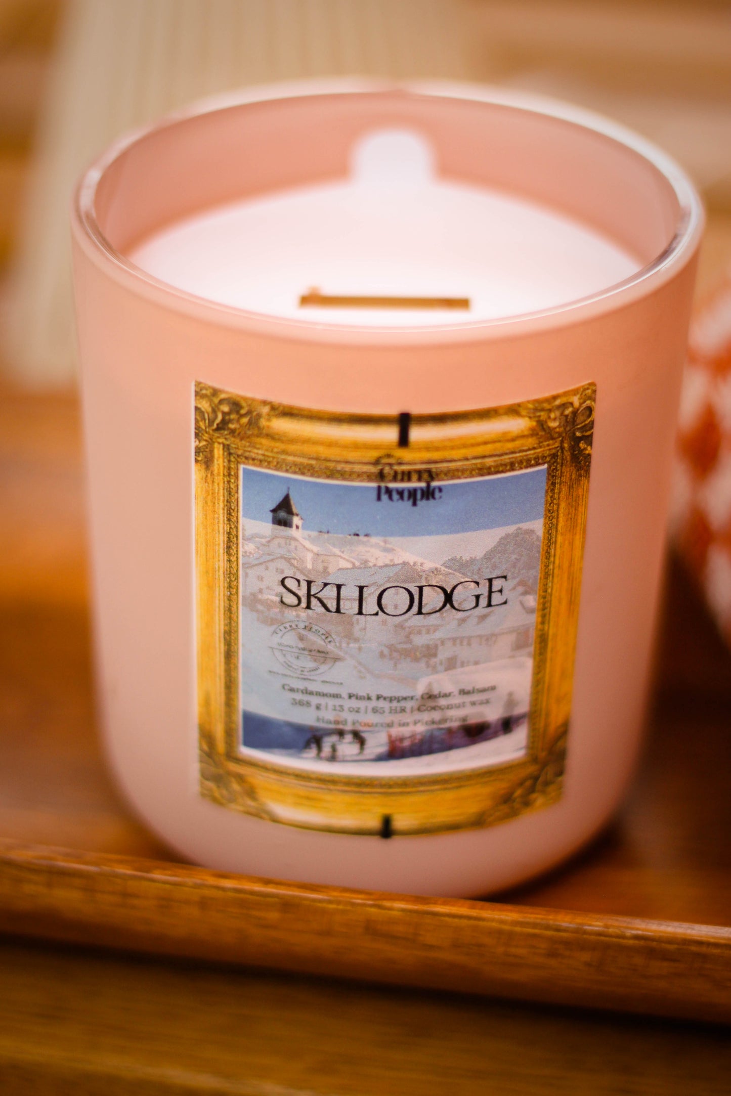 A pink glass jar candle labeled 'People Ski Lodge' sits on a wooden surface with a warm, softly blurred background.