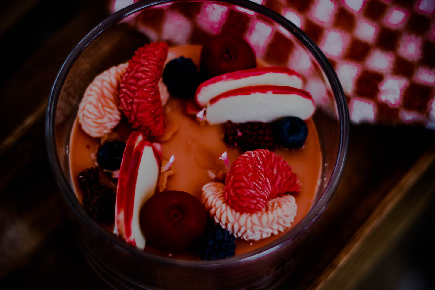 A dessert in a clear glass bowl, featuring a smooth orange-brown base topped with fresh raspberries, blackberries, blueberries, and white-and-red pastry slices, set on a dark wooden surface with a blurred red-and-white checkered cloth in the background.