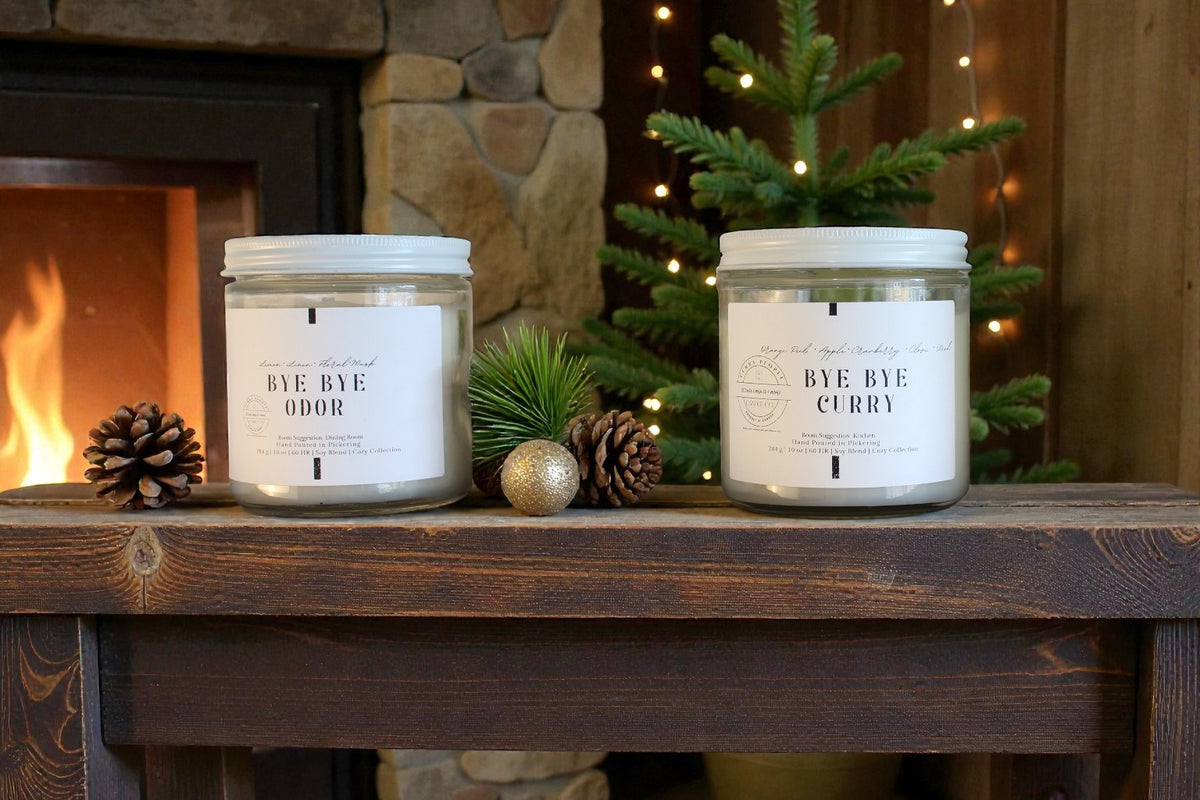 Two candles labeled 'Bye Bye Odor' and 'Bye Bye Curry' on a wooden surface with a fireplace and Christmas tree in the background.