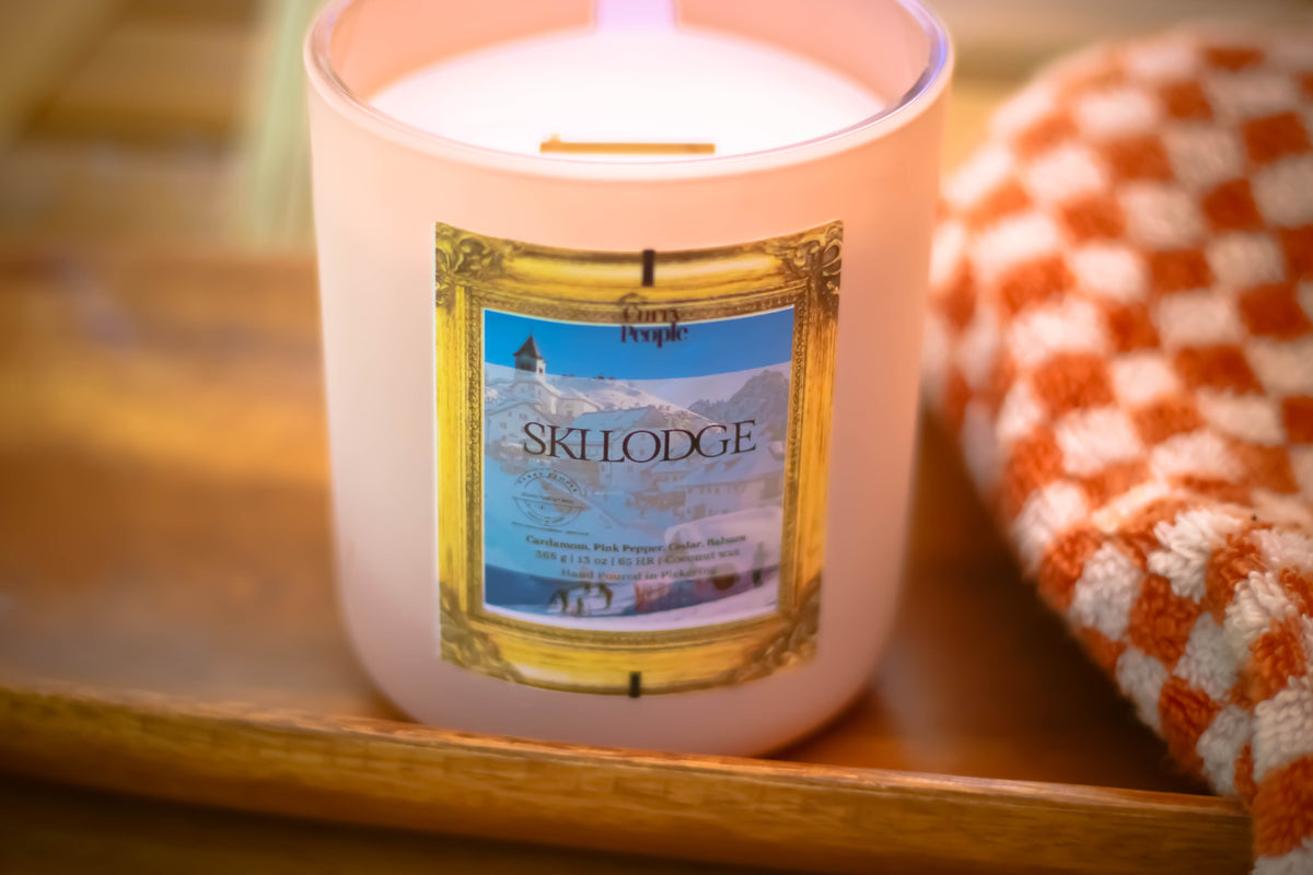 A lit scented candle in a frosted glass jar sits on a wooden tray beside a folded orange-and-white checkered towel. The front label reads 'Ski Lodge' with a mountain scene.