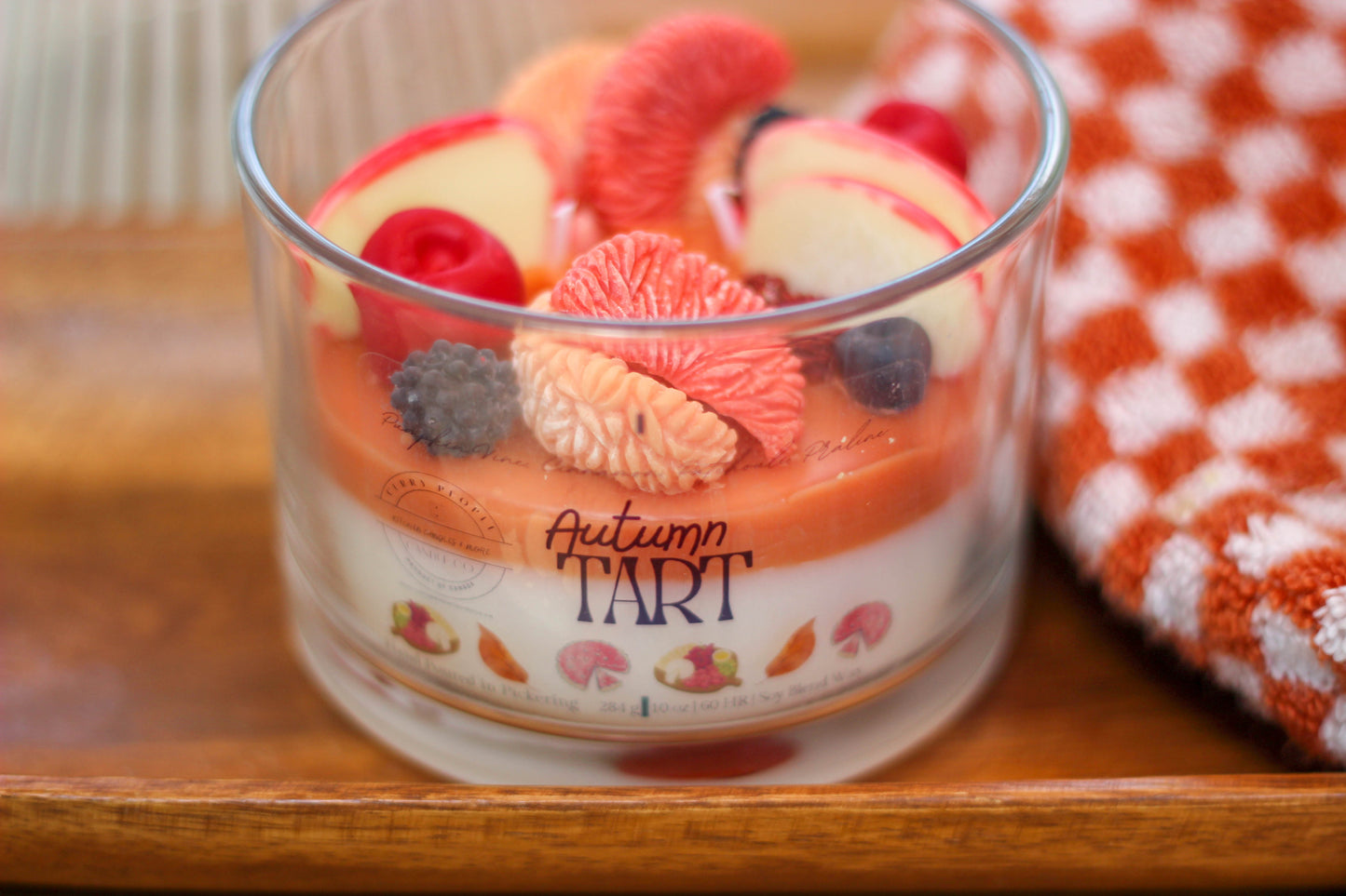 A glass jar candle labeled 'Autumn Tart' filled with layered orange and cream wax and topped with faux fruit slices, resting on a wooden surface next to an orange-and-white checkered cloth.