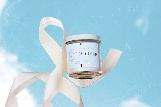 A clear glass jar soy candle labeled 'TEA CLOUD' with a white ribbon, against a blue sky with clouds.