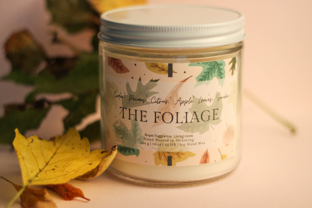 Candle jar labeled 'The Foliage' with autumn-themed design on a soft background.