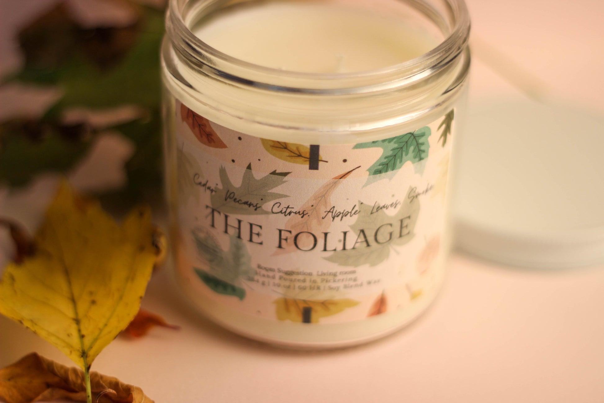 Candle jar labeled 'The Foliage' with autumn leaves on a neutral background