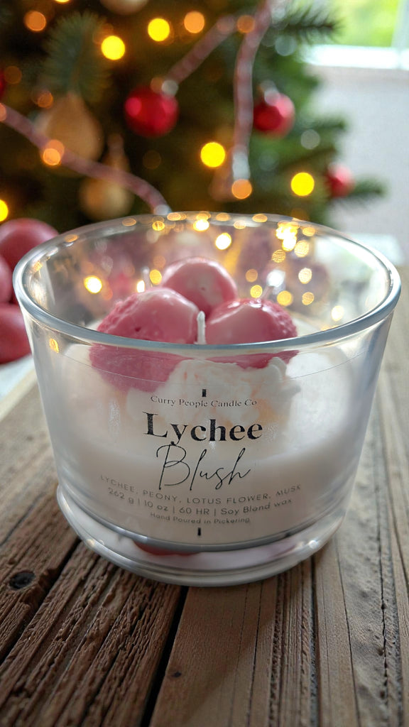 Lychee Blush candle with pink flowers in a glass container on a wooden surface with a blurred Christmas tree in the background.